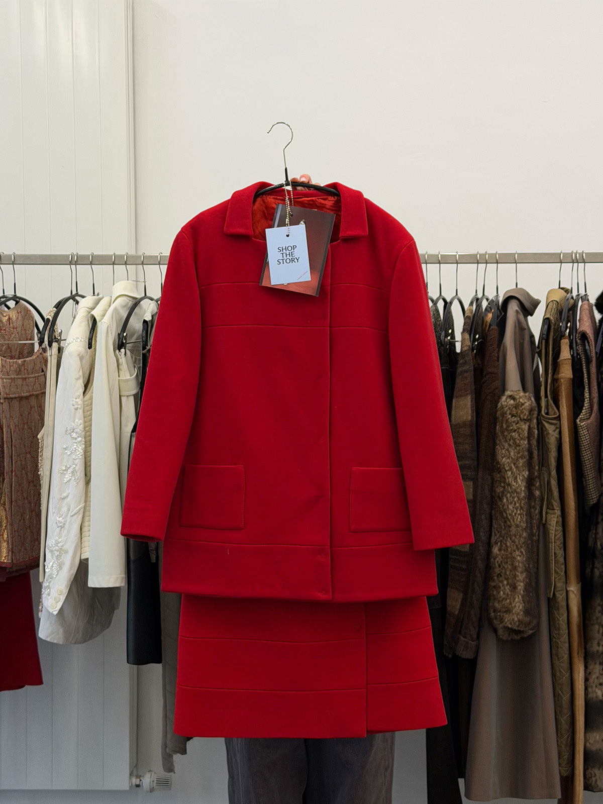 Red jacket and skirt in front of a clothing rack with various garments in a minimalistic room.