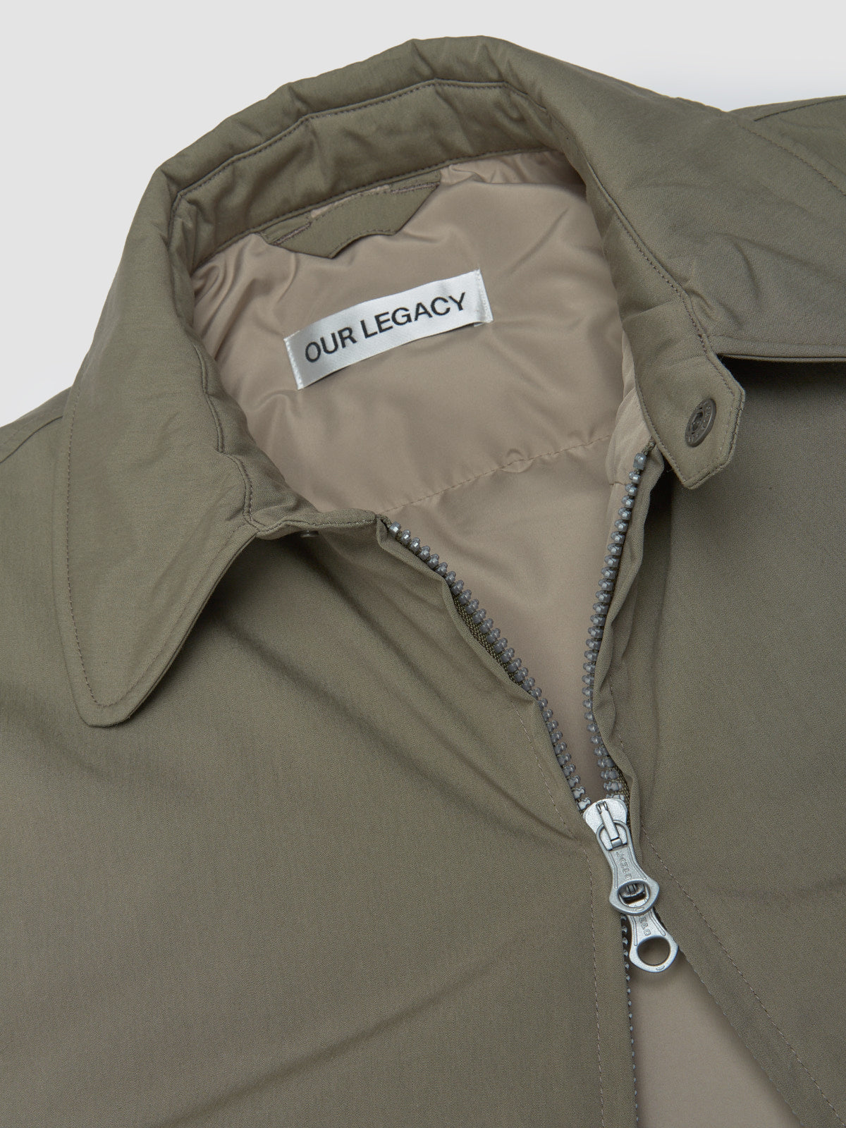 Close-up of a green jacket with a zipper and 'Our Legacy' label on a white background