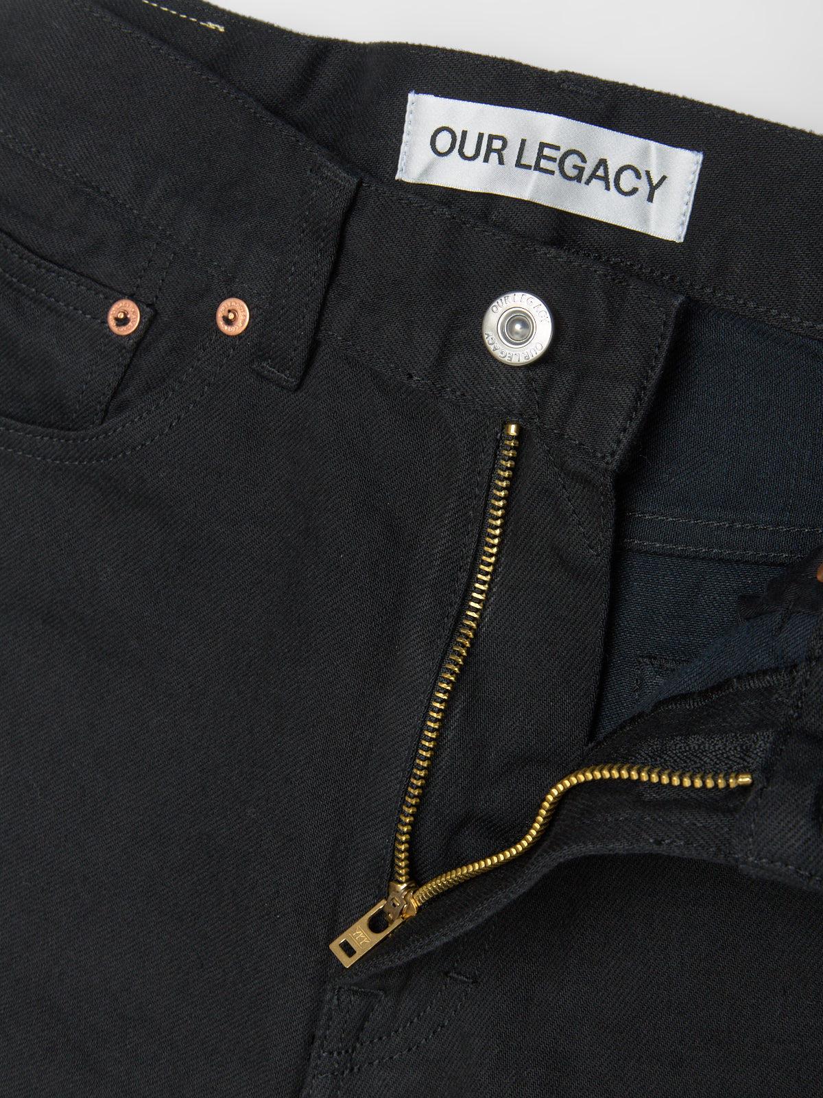 Close-up of black pants with a gold zipper and 'Our Legacy' label.