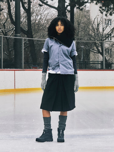 Person standing on an ice rink wearing a blue shirt, black skirt, and gray gloves.